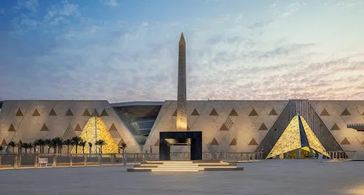 The Hanging Obelisk in the Grand Egyptian Museum plaza against the glass facade and illuminated golden triangles at sunset.