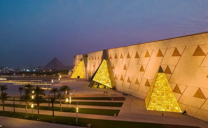 Illuminated Grand Egyptian Museum facade at sunset, featuring golden pyramidal designs with Giza Pyramids visible in the background.