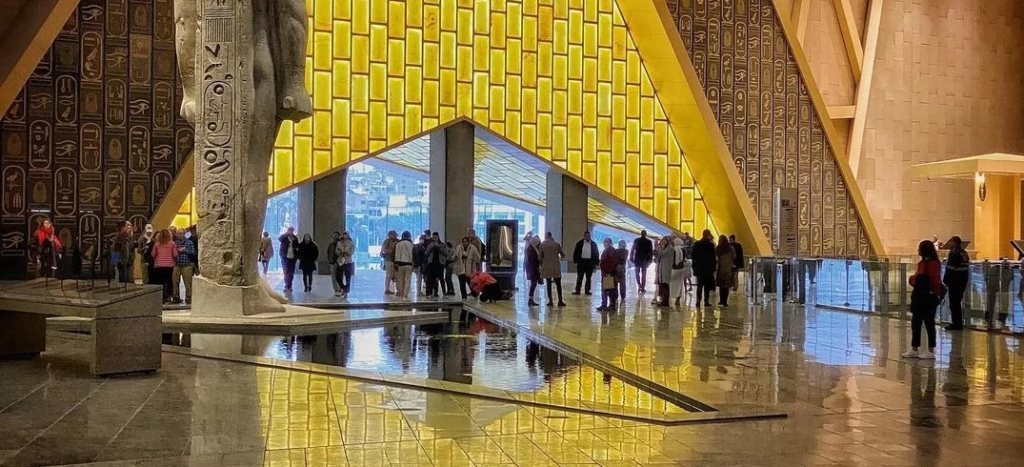Statue of Ramesses II in the Grand Egyptian Museum atrium with golden hieroglyphic walls and tourists reflecting on the floor.