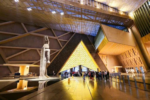 King Ramses II statue in the Grand Egyptian Museum atrium under modern geometric roofing and golden architectural lighting.