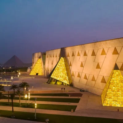Illuminated Grand Egyptian Museum facade at sunset, featuring golden pyramidal designs with Giza Pyramids visible in the background.